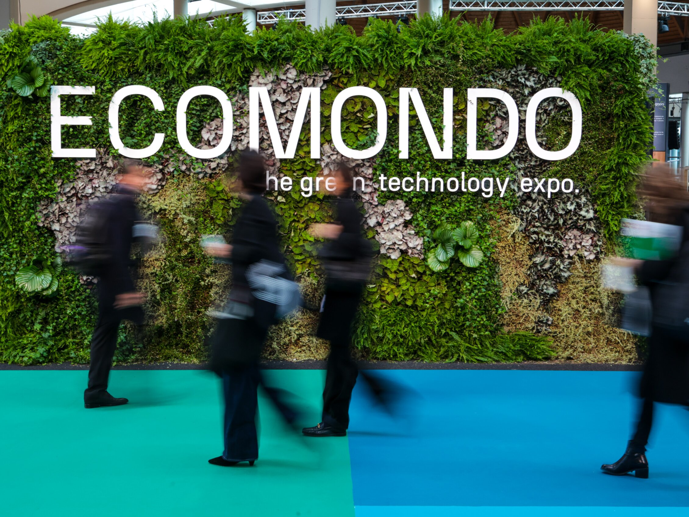 persone in movimento passano davanti ad un'installazione ricoperta di piante con la scritta ECOMONDO - the green technology expo